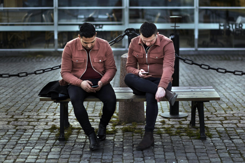 Swedish men are sitting on a bench and playing on the phones